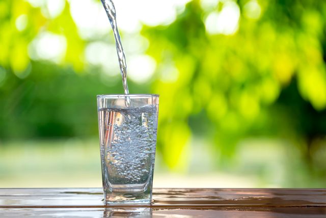 Pouring fresh water into a glass on a wooden table with a sunny, green outdoor background.