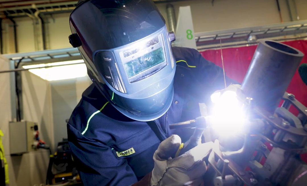 Welder wearing a helmet and safety gear, performing TIG welding on a metal pipe in an industrial workshop